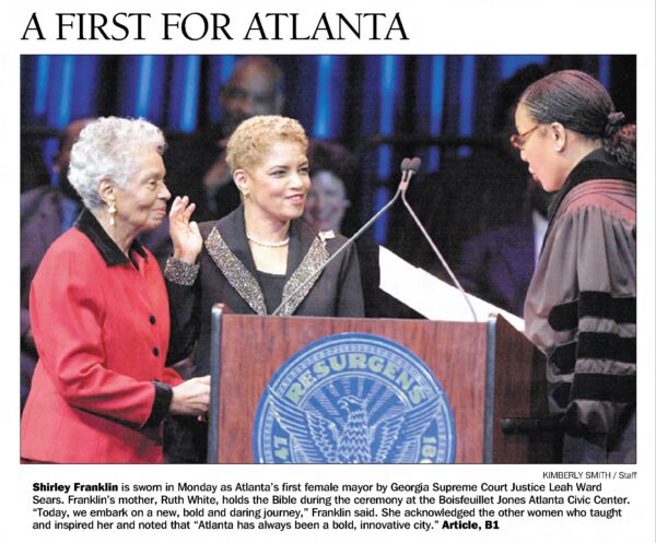 In a photo clipped from the newspaper an older Black woman with short gray hair wearing a red and black suit jacket stands next to a Black woman with short blonde hair wearing a dark suit with rhinestone accents and a pearl necklance holding her right hand in the air as she faces a Black female judge wearing robes.