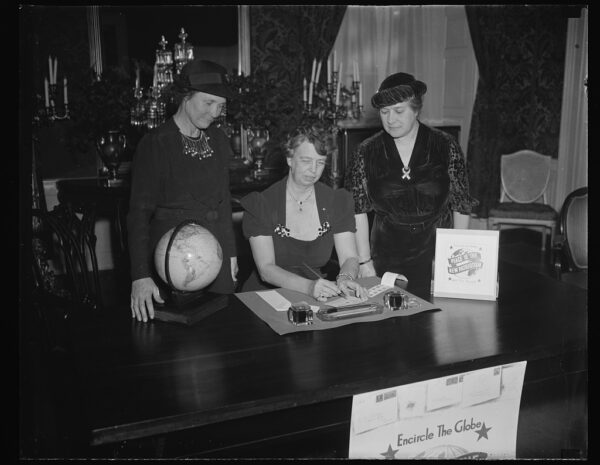 A posed photograph in black and white shows an elegant woman wearing a dress with large bejeweled accents and her pinned in a neat up-do seated at a desk writing a letter while two other women look on approvingly. Also on the desk are a globe and a small sign that reads 