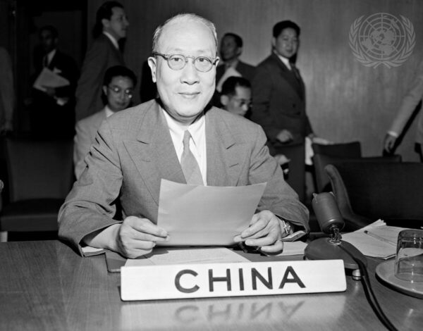 An older Asian man with smal oval glasses a small smile sits at a table looking directly into the camera while holding a piece of paper. In front of the man on the table is a microphone and a pacard that reads 