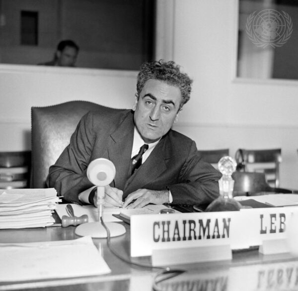 A middle-aged man with curly hair, cropped short on the sides and longer on top, and large dark eyebrows is sitting at a table behind a placard that reads 