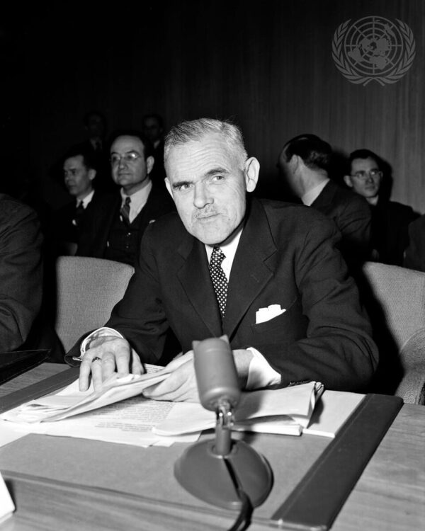 A caucasian man with partially grey hair and dark eyebrows wearing a dark suit with a polkadot bow tie looks slightly to the left of the photographer while sitting at a table in front of a microphone and holding a stack of messy papers.