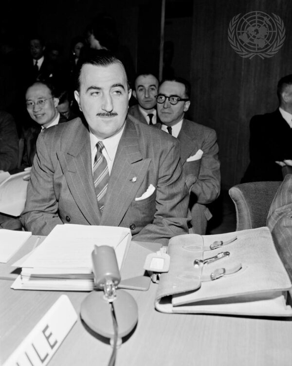 A hispanic middle-aged man with slicked-back dark hair and a thin dark mustache is seated at a table with a tall stack of papers, briefcase, and microphone in front of him.