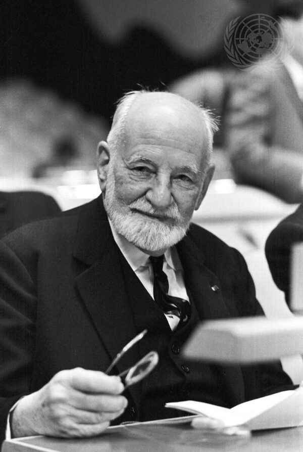 An elderly caucasian man with white hair and a short beard wearing a dark three suit sits at a table with a paper in front of him and a pair of glasses in his hand.