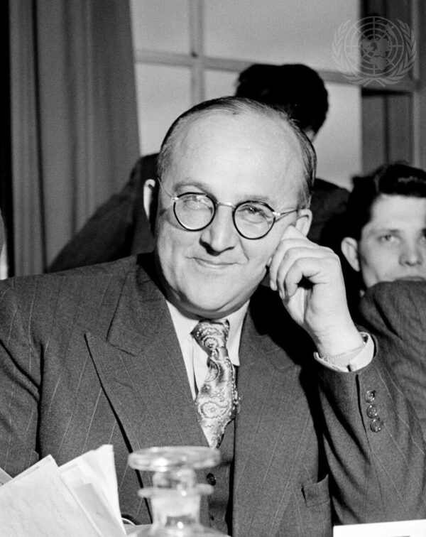 A black and white photo shows a seated caucasian man with round dark-rimmed glasses who is lightly resting his head on his hand while looking directly into the camera.