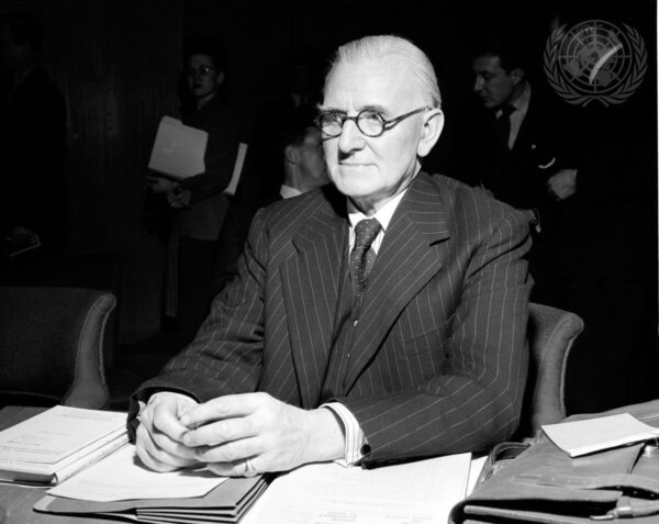 An elderly caucasian man with glasses and a pinstripe suit sits at a table with open books and stacks of papers in front of him.