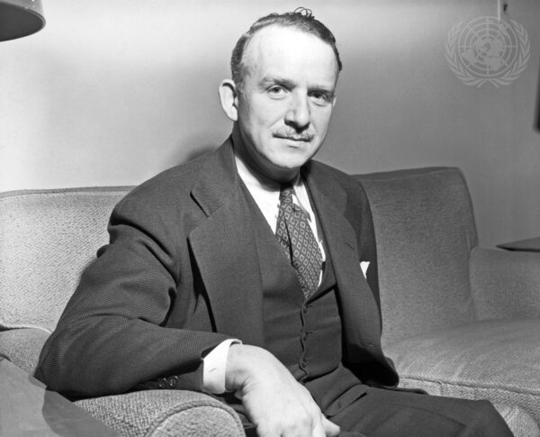 A middle-aged caucasian man wearing a dark three piece suit sits on a coach looking directly into the camera with a slight smile.