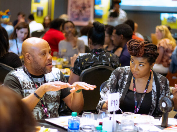 A bald Black man and a Black woman with braids are in discussion with others at a table.