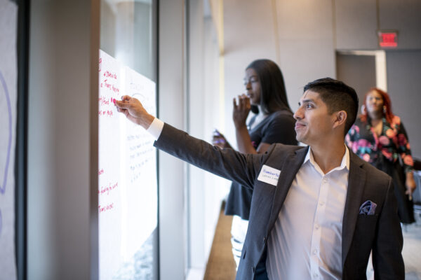 A male presenting person with dark hair and tan skin wearing a suit writes on a large sticky note on the wall as others look on.