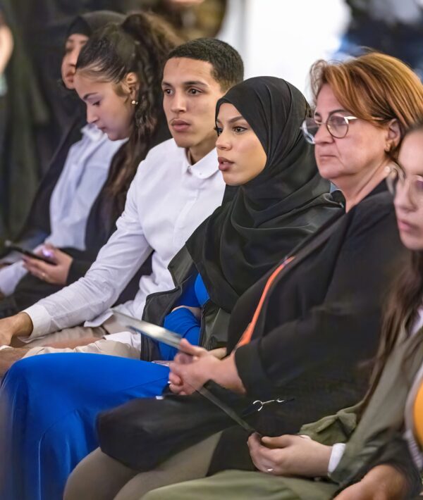 A row of attendees listens to a speaker. The guests central to the photo are a young woman wearing a black hijab and blue dress and a young man with close cropped dark hair wearing white button up shirt.