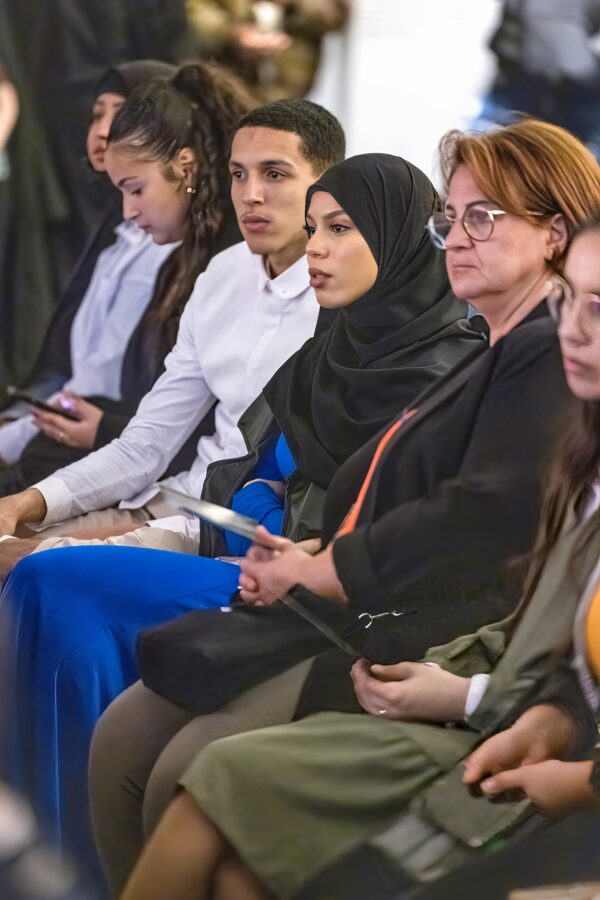 A row of attendees listens to a speaker. The guests central to the photo are a young woman wearing a black hijab and blue dress and a young man with close cropped dark hair wearing white button up shirt.