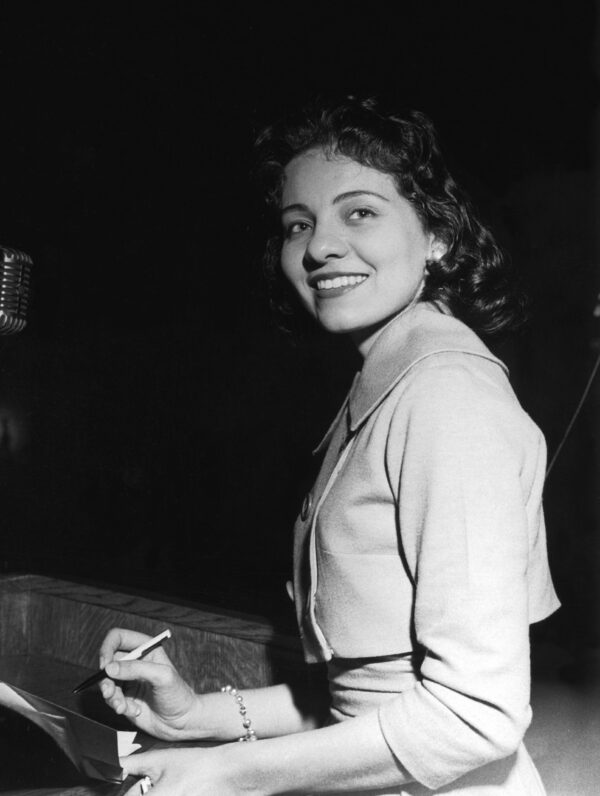 Candid photo of a young Black woman in a cropped suit jacket and matching skirt who is smiling widely while holding a pen in one hand and documents in the other.