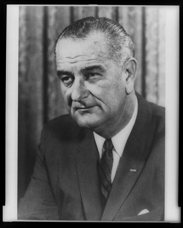 On older white man wearing a dark suit, white shirt, and dark tie poses with his hands clasped on a table and his gaze to the left of the photographer.