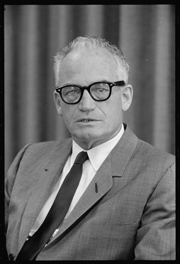 An older white man with short patchy white hair, wearing distinctive dark framed glasses, a gray suit, a dark tie, and white shirt.