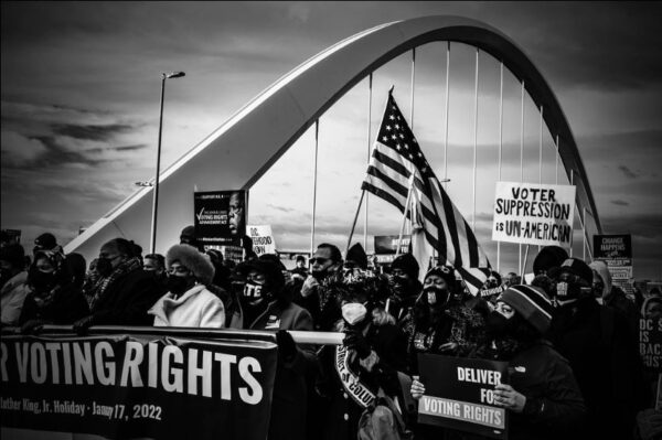 A black and white image of a voting rights protest. Attendees are wearing masks, holding signs, and one holds a large American flag at the center of the image.