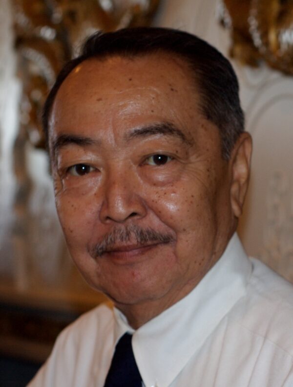 A portrait of an older Asian man with short dark hair and a mustache, wearing a white dress shirt and a dark tie. He is smiling slightly and looking directly at the camera. The background is softly lit and ornate, suggesting an elegant or formal setting.