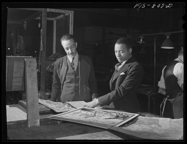 A black and white photo of two Black men in suits standing at a large table examining oversized documents. The back of a worker wearing a suit vest and apron is visible in the background.