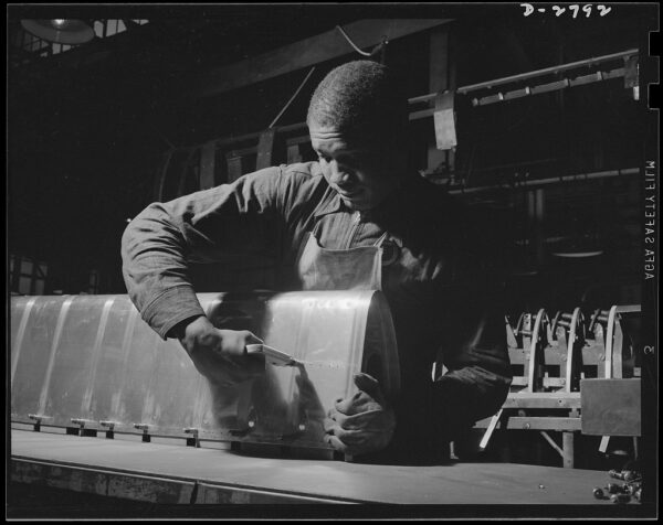Black man working in a factory, using a tool on large metal piece. Wearing long-sleeve shirt and apron. Industrial equipment in background. Black and white photo.
