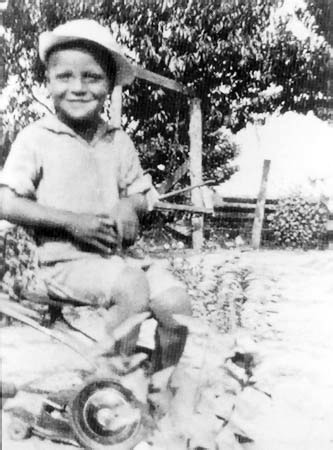 Small boy sitting on tricycle outdoors, wearing hat and casual clothing. Trees and wooden structures in background. Black and white photo.