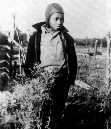 Young boy standing outdoors in rural setting, wearing jacket over shirt with hands in pockets. Wearing an aviator-style cap that fits snugly and covers the ears, similar to those worn by early 20th-century pilots. Plants, wooden structures, open fields, and trees in background. Black and white photo.
