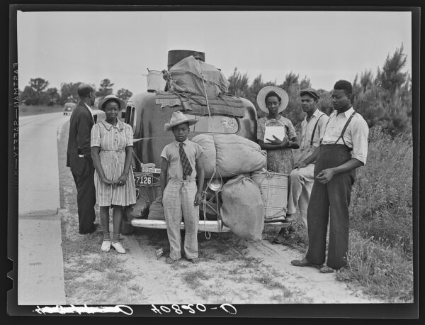 Old car on rural roadside, overloaded with bags and belongings. Girl in dress and hat stands in front; two boys and a girl at rear, one adjusting the load. Trees and bushes in background. Black and white photo.