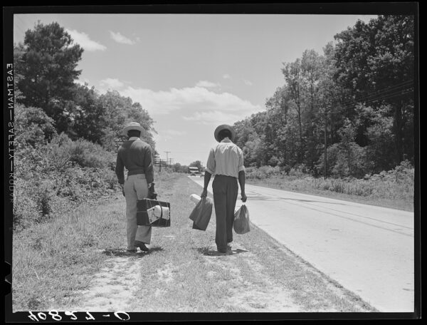 Two Black men walking along rural roadside, each carrying bags. Trees and bushes line the road. Black and white photo.