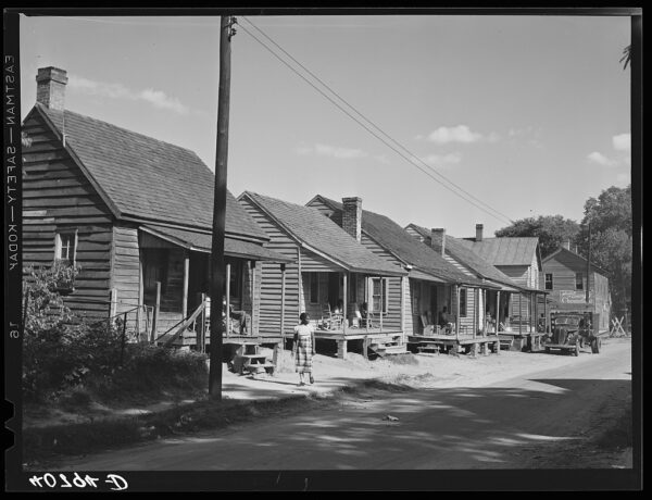 Row of wooden houses along dirt road. Black woman walking in front of the houses along the road. Power lines, utility pole, and old car visible. Porches have various items. Storefront in background. Black and white photo.
