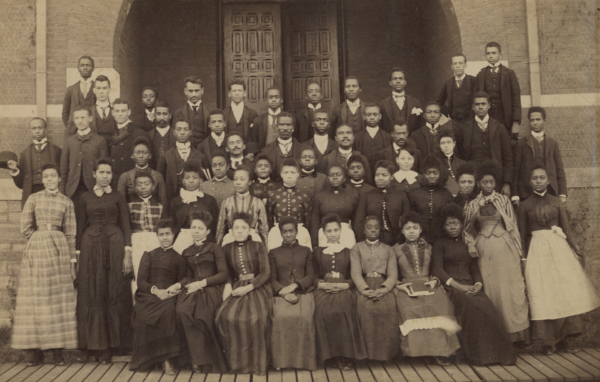 Black-and-white photo of a large group of students in formal clothes posed in front of a building.