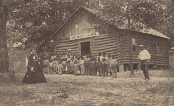 A sepia tone carte-de-visite of a Freedmen’s School. Standing closing together in front of a large building composed of logs and wide planks, several children are photographed on a bright day, surrounded by mature skinny trees. On the left of the photograph stands a woman wearing a large dark skirt and jacket. On the right of the photograph stands a man wearing hat, bright shirt, and dark trousers.