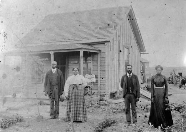 Four African American homesteaders standing in front of a small wooden house in Nicodemus, Kansas, including two men and two women dressed in formal clothing, with a porch and simple yard visible