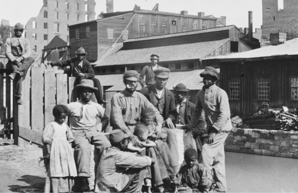 Group of newly freed African American men, women, and children gathered beside a canal in Richmond, Virginia, with wooden buildings and war-damaged structures in the background, 1865