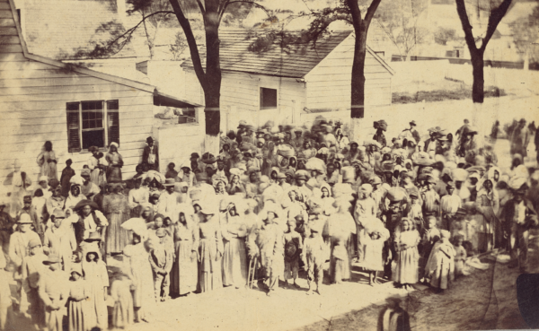 Large crowd of Black men, women, and children gathered outdoors near small wooden buildings on a South Carolina Plantation, late 19th century