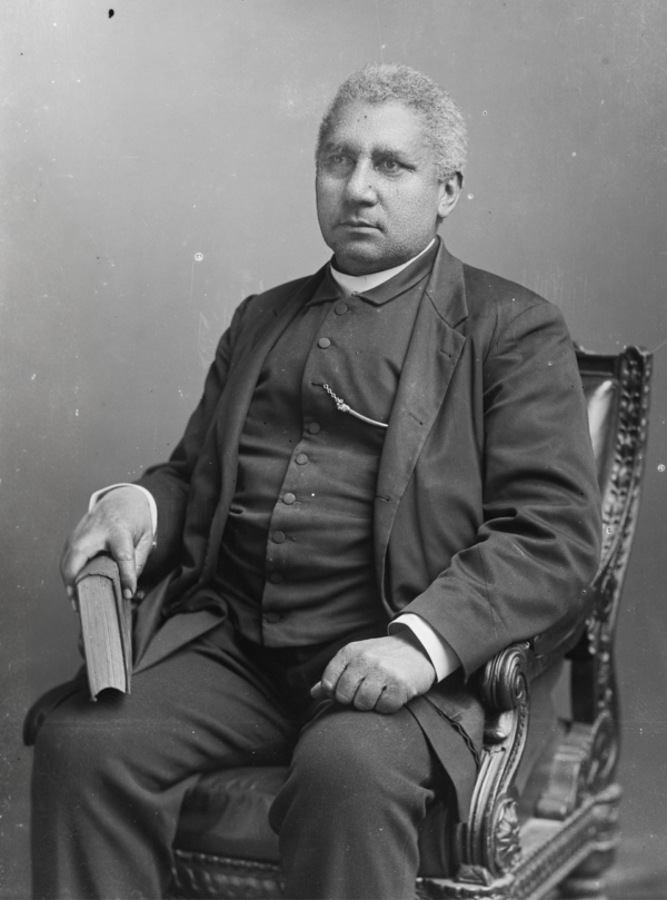 Portrait of Henry McNeal Turner seated in a chair, wearing a dark suit and holding a book, facing slightly to the side
