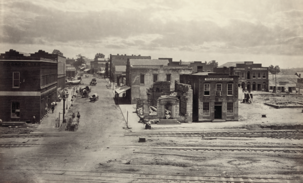 View up Peachtree Street in Atlanta from railroad tracks, showing damaged and partially destroyed buildings alongside intact storefronts, with wagons and pedestrians on the street after Union troops occupied the city in 1864