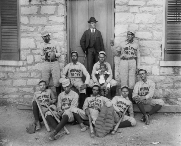 Group of African American baseball players from Morris Brown College posed with bats and catcher’s gear outside a stone building in Atlanta, with a well-dressed man standing behind them and a small child seated among the players