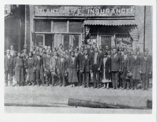 Large group of African American men and women in formal dress standing outside an Atlanta Life Insurance Company office, with storefront signs visible behind them