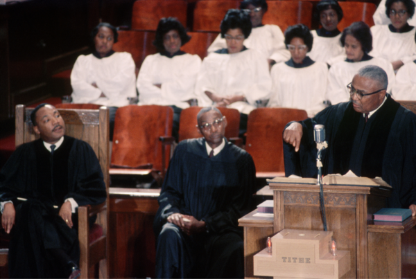 Martin Luther King Jr. seated in a church pulpit listening as Martin Luther King Sr. preaches at a lectern, with choir members seated behind them at Ebenezer Baptist Church in Atlanta