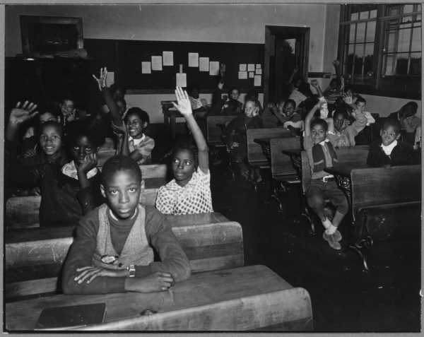 Black‑and‑white photograph of a classroom filled with young students seated at wooden desks. Many students have one arm raised, as if responding to a question. In the foreground, one student sits at a desk facing the camera while others behind and to the sides raise their hands. The room includes a chalkboard or bulletin board along the back wall with several papers pinned up, a doorway at center-right, and large windows on the right side letting in light. Books and classroom materials are stacked near some desks, and the overall setting suggests an older, traditional schoolroom.