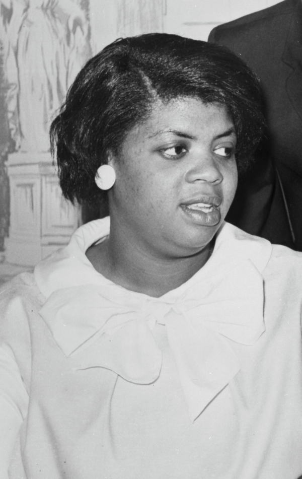 A candid black and white photograph of a Black woman with a short straight bob, large round earrings, and a light-colored jacket with a wide collar.