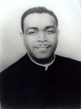 Black and white portrait of an African American man with short hair and a thing moustache wearing a priest's collar and smiling at the camera.