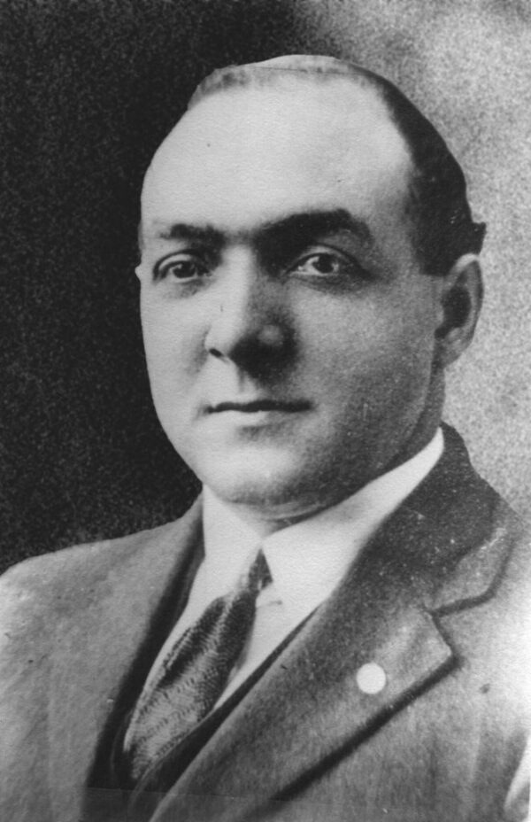 Black and white photograph of a middle-aged man with light skin and a receding hairline, wearing a suit and tie, looks into the camera and smiles slightly.