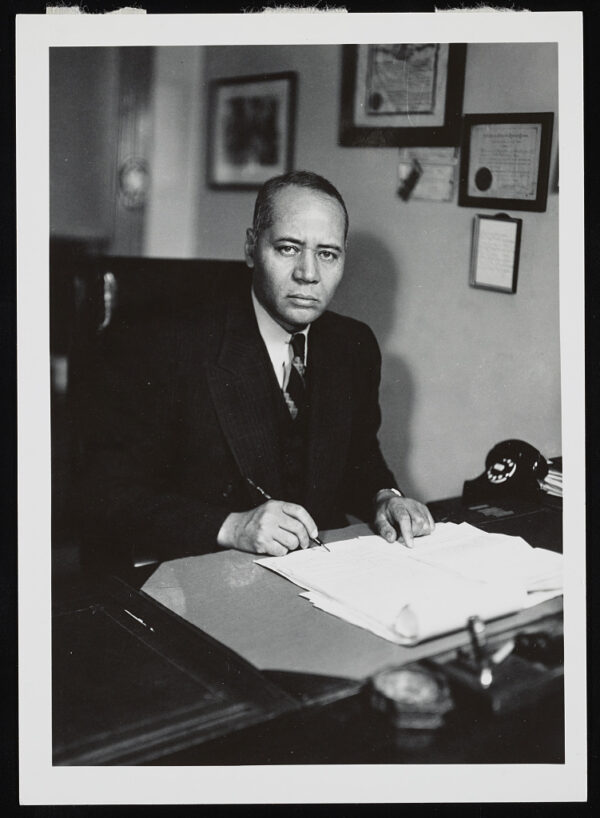 An older man wearing a suit and tie sits at a desk with a stack of papers in front of him and a pen in his hand looks sternly into the camera.