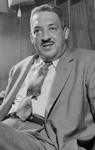 A black and white image of a middle aged man with a moustache wearing a suite and tie speaking to someone off camera.