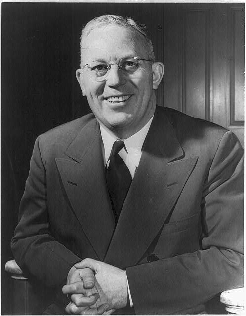 An older white man smiles widely at the camera. He wears thin metal-rimmed glasses and a suit and tie.