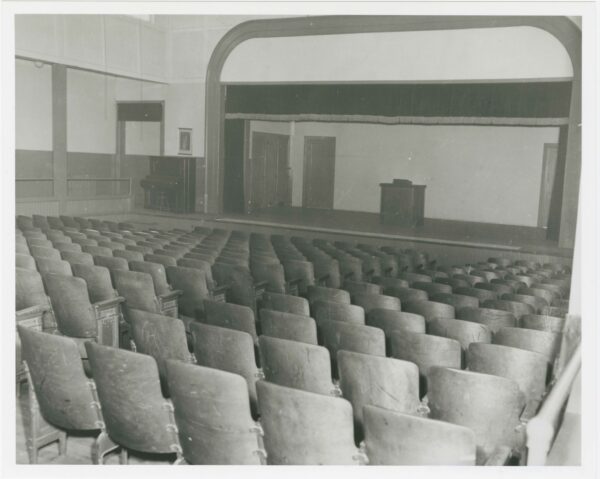 Photo of an auditorium showing rows of built-in seating and a stage with a curtain, podium, and a piano.